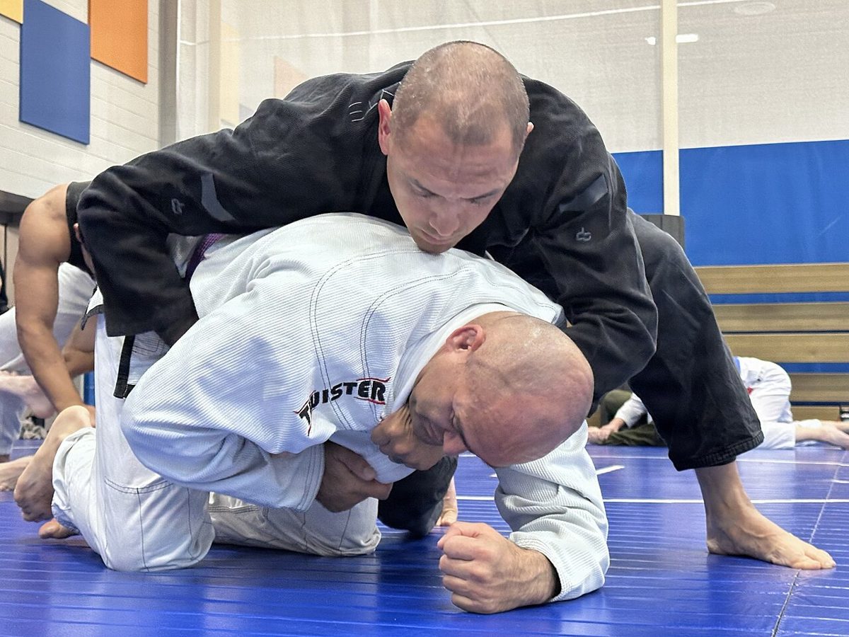 Praticante de kimono branco treinando no chão com o instrutor de kimono preto durante uma aula de jiu-jitsu