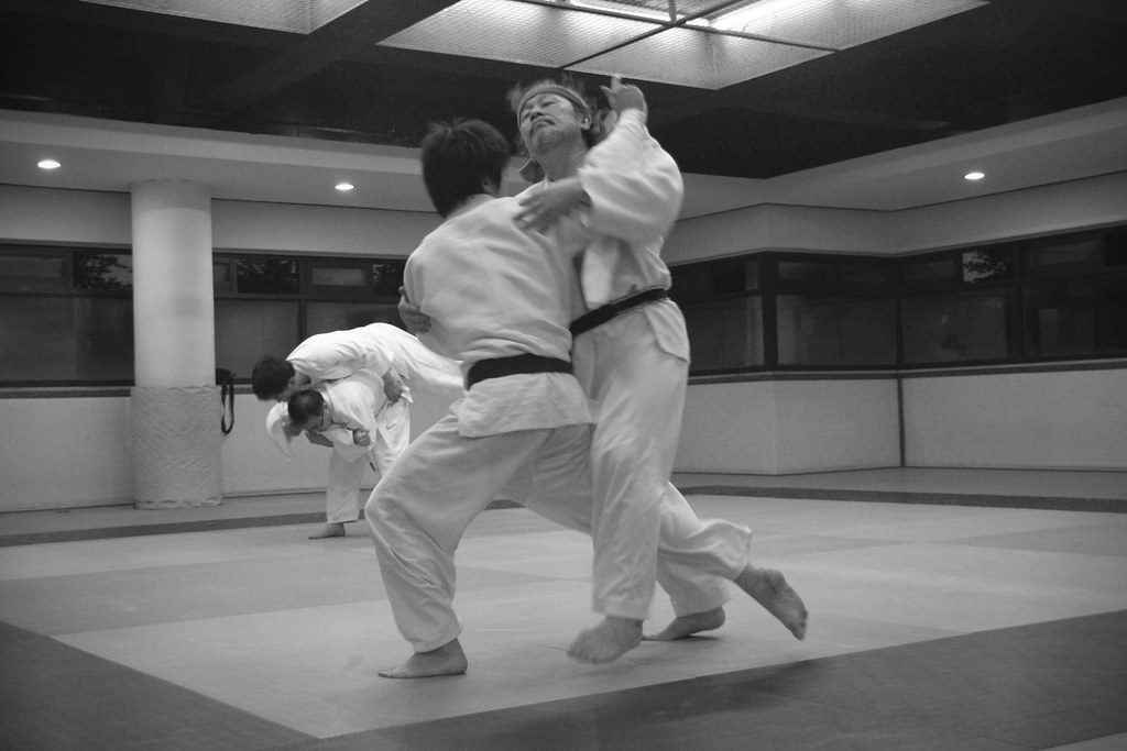 Foto em preto e branco de dois praticantes de kimono repetindo a entrada de uma queda numa academia