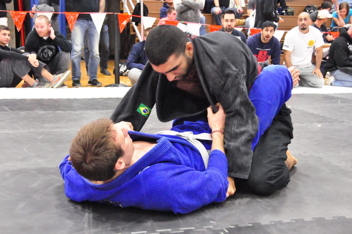Atleta de kimono azul segurando o adversário de kimono preto na guarda fechada durante um torneio