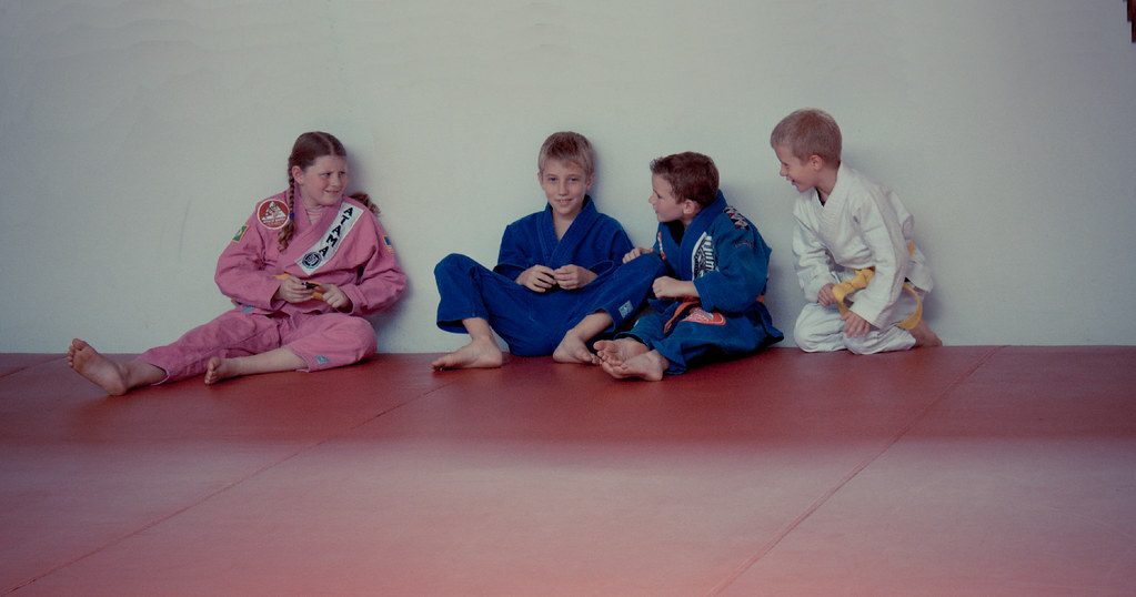 Quatro crianças de kimono sentadas no tatame encostadas na parede de uma academia