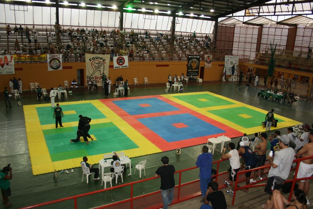 Vista do alto de um ginásio com tatames coloridos e várias lutas de jiu-jitsu acontecendo ao mesmo tempo